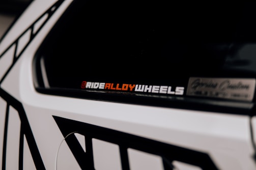 A close-up of a car accent showcasing a decal or sticker that reads 'RIDEALLOYWHEELS' in a bold font. Part of the vehicle is visible with contrasting black and white geometric patterns on the body.