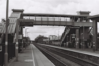 A deserted train station platform with a large steel pedestrian bridge overhead, multiple staircases leading up to the bridge, and various lampposts and signage along the platform. The tracks run parallel between the platforms, extending into the distance.