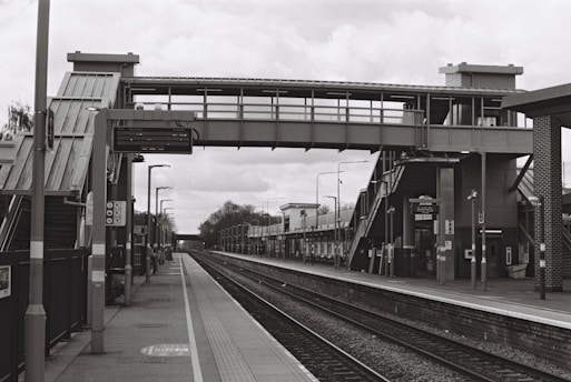 A deserted train station platform with a large steel pedestrian bridge overhead, multiple staircases leading up to the bridge, and various lampposts and signage along the platform. The tracks run parallel between the platforms, extending into the distance.