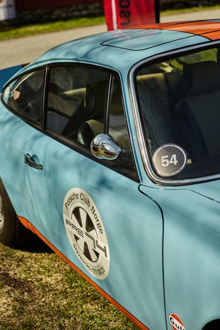 A vintage blue car with an orange stripe and the logo of Porsche Club Norge is parked on grass. The car door features a circular decal with the text 'Porsche Club Norge.' A racing number '54' is displayed on the side window. A red sign and some greenery are visible in the background.