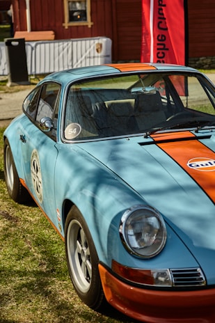 A vintage sports car is parked outdoors on a grassy area. The car has a light blue body with an orange racing stripe down the center. It features classic round headlights and is adorned with racing decals, including the number 54 and a Gulf logo. A red vertical banner and a wooden building are visible in the background.