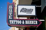A street sign for a business named Elite Tattoo & Barber with vintage-style lettering, prominently featuring an arrow. An additional sign above shows another business name in a modern script.