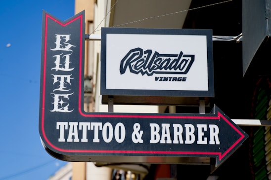 A street sign for a business named Elite Tattoo & Barber with vintage-style lettering, prominently featuring an arrow. An additional sign above shows another business name in a modern script.