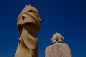 Two sculptural structures with distinctive, artistic forms against a clear blue sky. The larger structure on the left has smooth, flowing curves with a modern abstract design, while the one on the right is composed of smaller stone fragments, creating a textured surface.