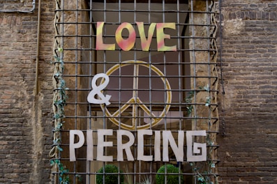 A storefront window is covered with metal bars, adorned with vines. The words 'LOVE', '&', and 'PIERCING' are mounted on the bars, with a large peace symbol in the background. The brick wall surrounds the window, adding a rustic feel.