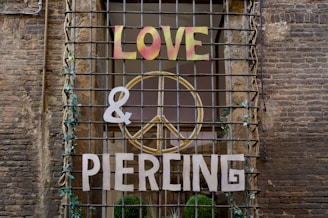 A storefront window is covered with metal bars, adorned with vines. The words 'LOVE', '&', and 'PIERCING' are mounted on the bars, with a large peace symbol in the background. The brick wall surrounds the window, adding a rustic feel.