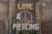 A storefront window is covered with metal bars, adorned with vines. The words 'LOVE', '&', and 'PIERCING' are mounted on the bars, with a large peace symbol in the background. The brick wall surrounds the window, adding a rustic feel.