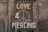 A storefront window is covered with metal bars, adorned with vines. The words 'LOVE', '&', and 'PIERCING' are mounted on the bars, with a large peace symbol in the background. The brick wall surrounds the window, adding a rustic feel.