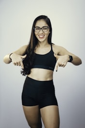 A woman with long dark hair wearing glasses, a black sports bra, and black shorts is smiling and pointing downwards with both hands. She is also wearing a smartwatch on her wrist, and her nails are painted black.