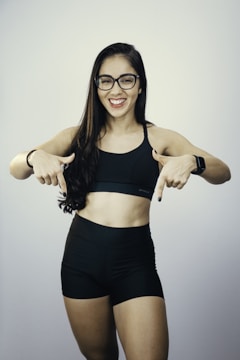 A woman with long dark hair wearing glasses, a black sports bra, and black shorts is smiling and pointing downwards with both hands. She is also wearing a smartwatch on her wrist, and her nails are painted black.