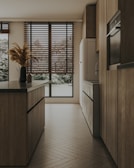 Modern kitchen featuring warm-toned laminate flooring and stylish roller blinds on large windows.