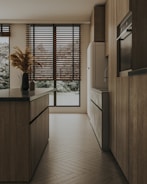 A modern kitchen with sleek wooden cabinets and an island featuring dark countertops. Large windows with wooden blinds allow natural light to filter in, highlighting the warm tones of the wooden floor. A simple arrangement of pampas grass in a vase adds a touch of natural decor.