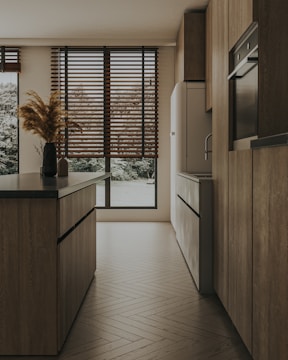 A modern kitchen with sleek wooden cabinets and an island featuring dark countertops. Large windows with wooden blinds allow natural light to filter in, highlighting the warm tones of the wooden floor. A simple arrangement of pampas grass in a vase adds a touch of natural decor.