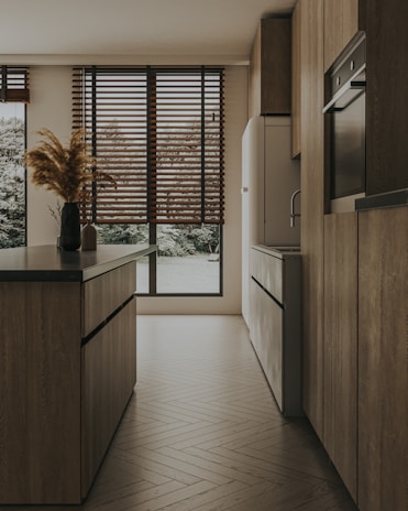 A modern kitchen with sleek wooden cabinets and an island featuring dark countertops. Large windows with wooden blinds allow natural light to filter in, highlighting the warm tones of the wooden floor. A simple arrangement of pampas grass in a vase adds a touch of natural decor.