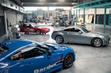 A spacious automotive workshop with polished concrete floors is shown. Three cars, a red one, a blue one, and a silver one, are parked indoors. The background reveals various mechanical equipment and scattered boxes, suggesting ongoing work.