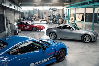 A spacious automotive workshop with polished concrete floors is shown. Three cars, a red one, a blue one, and a silver one, are parked indoors. The background reveals various mechanical equipment and scattered boxes, suggesting ongoing work.