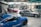 A spacious automotive workshop with polished concrete floors is shown. Three cars, a red one, a blue one, and a silver one, are parked indoors. The background reveals various mechanical equipment and scattered boxes, suggesting ongoing work.