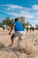 A group of people are engaging in physical training on a sandy beach. A person in the foreground is in motion, kicking up sand as they run or exercise. Several palm trees and a tall building are visible in the background under a clear blue sky.