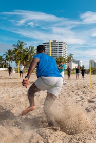 A group of people are engaging in physical training on a sandy beach. A person in the foreground is in motion, kicking up sand as they run or exercise. Several palm trees and a tall building are visible in the background under a clear blue sky.