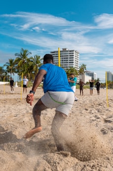A group of people are engaging in physical training on a sandy beach. A person in the foreground is in motion, kicking up sand as they run or exercise. Several palm trees and a tall building are visible in the background under a clear blue sky.