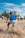 A group of people are engaging in physical training on a sandy beach. A person in the foreground is in motion, kicking up sand as they run or exercise. Several palm trees and a tall building are visible in the background under a clear blue sky.