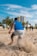 A group of people are engaging in physical training on a sandy beach. A person in the foreground is in motion, kicking up sand as they run or exercise. Several palm trees and a tall building are visible in the background under a clear blue sky.