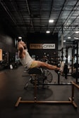 A person is exercising on a hyperextension bench in a gym with rows of equipment visible in the background and a large sign displaying 'PER4ORM'. The gym is dimly lit with overhead lights and a high ceiling.