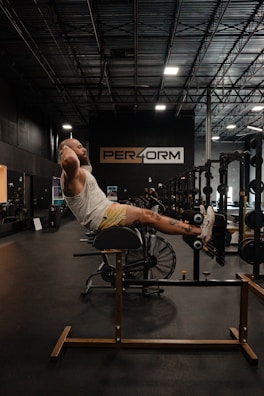 A person is exercising on a hyperextension bench in a gym with rows of equipment visible in the background and a large sign displaying 'PER4ORM'. The gym is dimly lit with overhead lights and a high ceiling.
