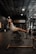 A person is exercising on a hyperextension bench in a gym with rows of equipment visible in the background and a large sign displaying 'PER4ORM'. The gym is dimly lit with overhead lights and a high ceiling.