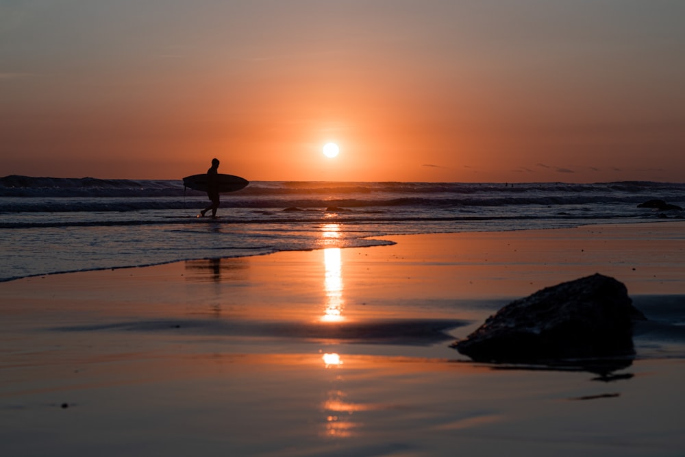 eine person, die ein surfbrett an einem strand bei sonnenuntergang hält