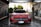 A red Mercedes-Benz sports car is in a car wash, covered in soap suds. The setting appears to be an outdoor car washing bay with advertisements and equipment visible on either side. The background includes some greenery and a building.