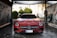 A red Mercedes-Benz sports car is in a car wash, covered in soap suds. The setting appears to be an outdoor car washing bay with advertisements and equipment visible on either side. The background includes some greenery and a building.