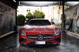 A red Mercedes-Benz sports car is in a car wash, covered in soap suds. The setting appears to be an outdoor car washing bay with advertisements and equipment visible on either side. The background includes some greenery and a building.