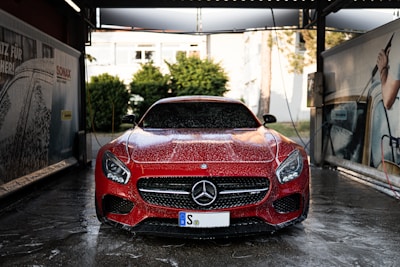 A red Mercedes-Benz sports car is in a car wash, covered in soap suds. The setting appears to be an outdoor car washing bay with advertisements and equipment visible on either side. The background includes some greenery and a building.