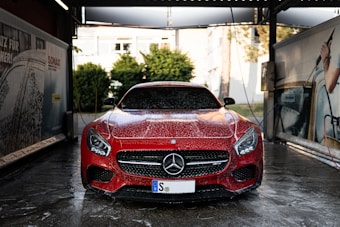 A red Mercedes-Benz sports car is in a car wash, covered in soap suds. The setting appears to be an outdoor car washing bay with advertisements and equipment visible on either side. The background includes some greenery and a building.