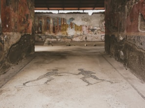 An ancient mosaic floor features two human figures, possibly athletes, engaging in an athletic activity. The walls surrounding the mosaic are constructed with old stone and faded frescoes, suggesting historical significance. The background includes more remnants of painted murals and architectural ruins.