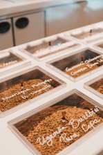 A series of transparent containers filled with coffee beans are neatly arranged on a surface. Each container has a label written in white script, indicating the origin or type of coffee beans inside. The background is softly out of focus, showing part of a kitchen setup.