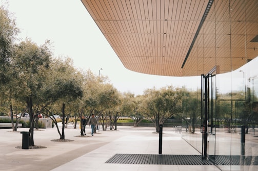 A modern architectural scene featuring a large glass building with a wooden ceiling overhang. The exterior reflects a row of neatly arranged trees lining a spacious concrete walkway. A couple of people are seen walking near the entrance, adding a sense of movement to the tranquil setting.
