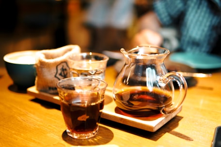 A rustic wooden table with an open box of assorted coffee packs and a steaming cup of coffee beside it