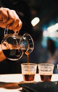 A hand is pouring dark coffee from a glass pitcher into small glass cups, with a warm and inviting ambiance created by blurred background lights.