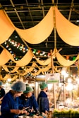 A vibrant market scene with people wearing blue uniforms and hair nets, busy preparing or serving food. Yellow fabric banners hang from above, and colorful string lights add a festive touch. A sign reading 'BBQ' is visible, and the atmosphere seems lively and bustling.