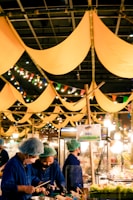 A vibrant market scene with people wearing blue uniforms and hair nets, busy preparing or serving food. Yellow fabric banners hang from above, and colorful string lights add a festive touch. A sign reading 'BBQ' is visible, and the atmosphere seems lively and bustling.