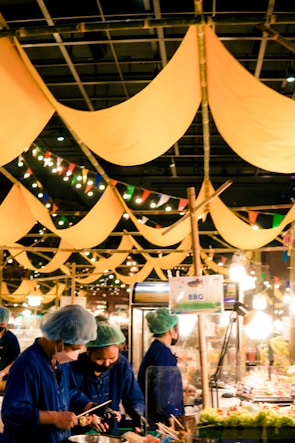 A vibrant market scene with people wearing blue uniforms and hair nets, busy preparing or serving food. Yellow fabric banners hang from above, and colorful string lights add a festive touch. A sign reading 'BBQ' is visible, and the atmosphere seems lively and bustling.