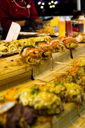A variety of sushi rolls arranged on bamboo mats. The sushi appears to be topped with various ingredients such as salmon and mayonnaise. In the background, there are condiments in squeeze bottles and a person in a red uniform preparing food.