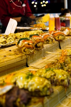 A variety of sushi rolls arranged on bamboo mats. The sushi appears to be topped with various ingredients such as salmon and mayonnaise. In the background, there are condiments in squeeze bottles and a person in a red uniform preparing food.