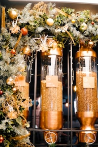 Three large clear cylindrical containers are filled with coffee beans and are mounted in a metallic frame. Labels on each container read 'Early Yellow', 'Green Beans', and 'First Crack'. The containers are adorned with a decorative garland featuring lush green foliage, golden elements, and ornaments in tones of gold and red.