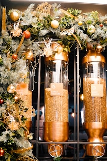 Three large clear cylindrical containers are filled with coffee beans and are mounted in a metallic frame. Labels on each container read 'Early Yellow', 'Green Beans', and 'First Crack'. The containers are adorned with a decorative garland featuring lush green foliage, golden elements, and ornaments in tones of gold and red.