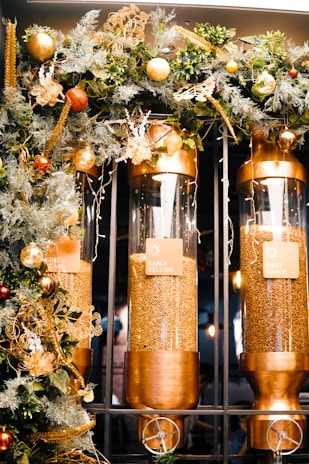Three large clear cylindrical containers are filled with coffee beans and are mounted in a metallic frame. Labels on each container read 'Early Yellow', 'Green Beans', and 'First Crack'. The containers are adorned with a decorative garland featuring lush green foliage, golden elements, and ornaments in tones of gold and red.