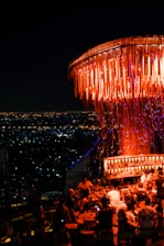 A vibrant rooftop bar filled with people enjoying drinks in a lively atmosphere. The setting is adorned with an elaborate light fixture casting a warm orange glow. The cityscape in the background shows a sea of sparkling lights under a dark night sky.