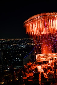 A vibrant rooftop bar filled with people enjoying drinks in a lively atmosphere. The setting is adorned with an elaborate light fixture casting a warm orange glow. The cityscape in the background shows a sea of sparkling lights under a dark night sky.